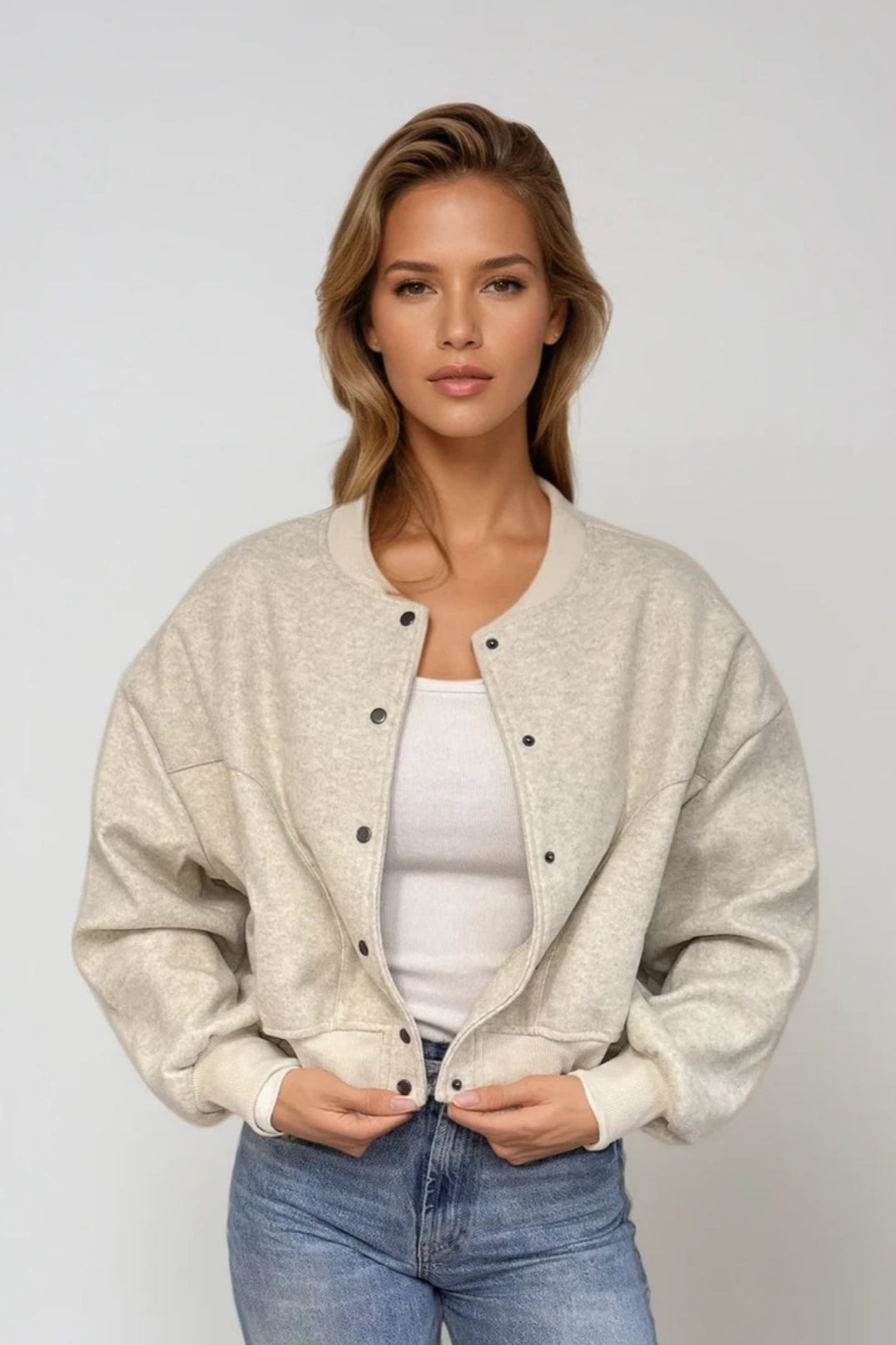A female model wearing a beige bomber jacket with a round neckline and long sleeves, paired with a white top and blue jeans. Accessories include a belt and white shoes.
