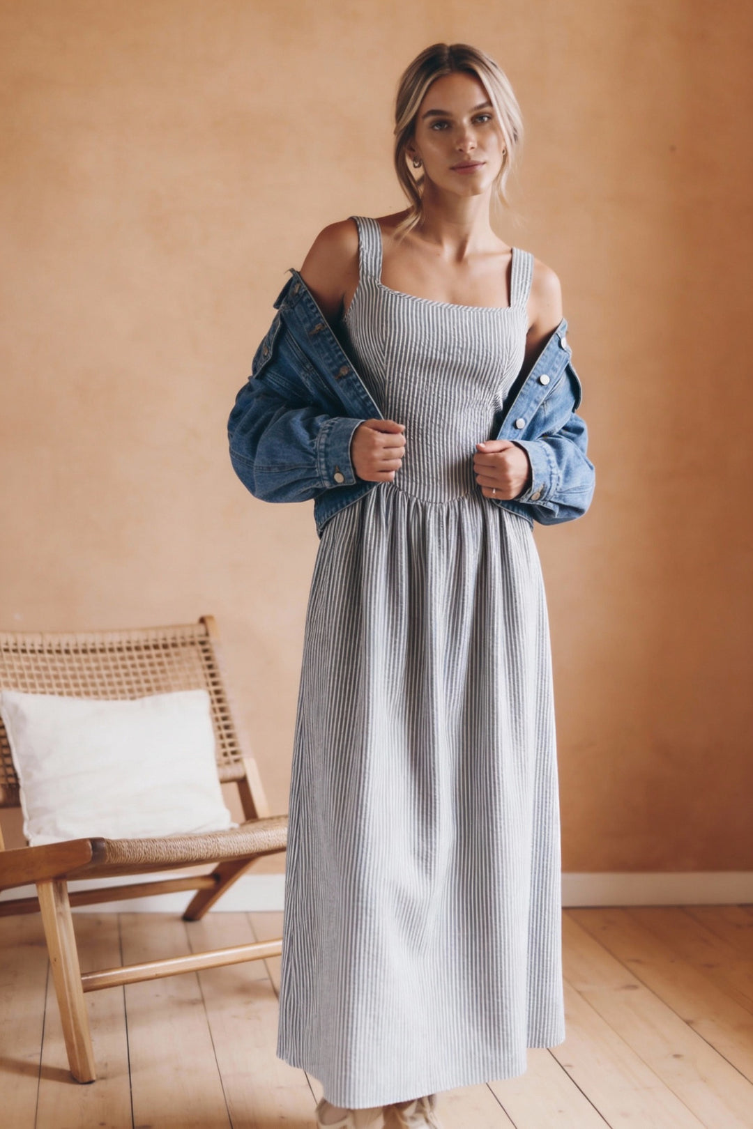 Woman wearing a light blue dress with a denim jacket in a room with wooden flooring and a wicker chair.