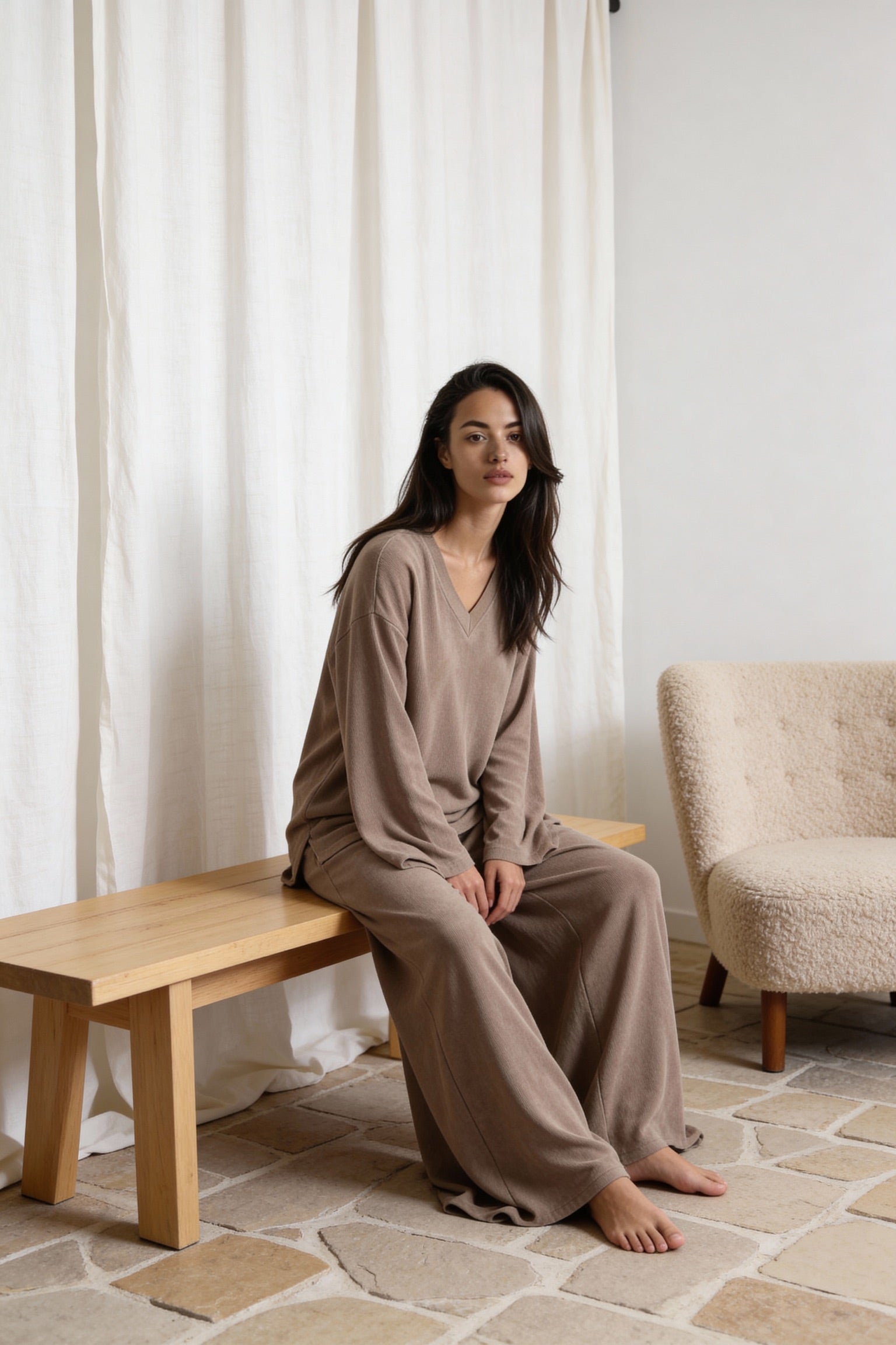 Woman sitting on a wooden bench in a room with white curtains and a beige armchair. Wearing the Terry & co set by priv