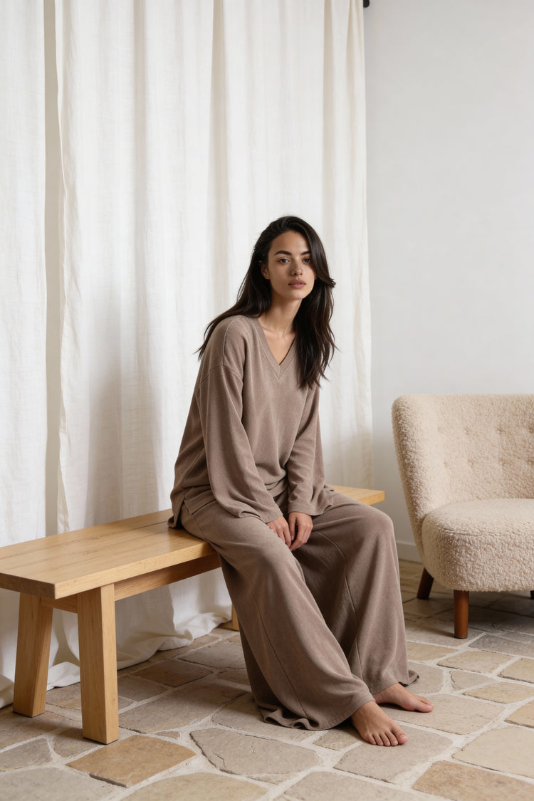Woman sitting on a wooden bench in a room with white curtains and a beige armchair. Wearing the Terry & co set by priv